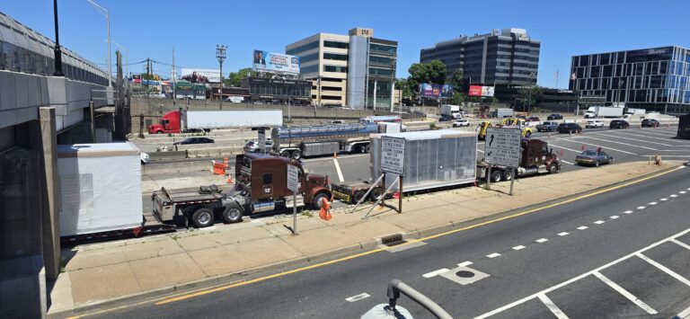 Heavy Haul Solutions equipment parked at a staging yard, including a red semi tractor with enclosed trailer and additional trailers nearby, with city buildings in background.