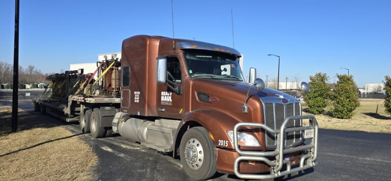 Heavy Haul Solutions semi tractor with step deck trailer parked in yard with industrial transformers in background.