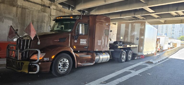 Semi tractors with Heavy Haul Solutions branding parked under bridge with modular units on lowboy trailers in urban setting.