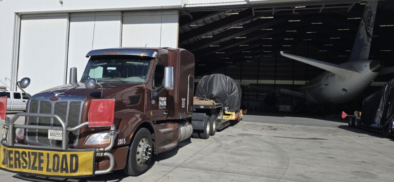 Semi tractor with oversize load placard parked in at an airport near hangar with loaded trailer in background.