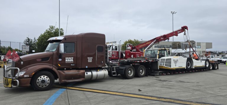 Semi tractor with step deck trailer parked in yard next to red industrial crane and white heavy-haul equipment.