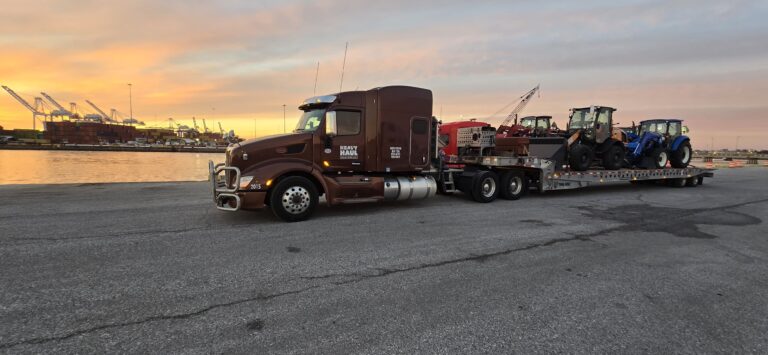 Tractor-trailer rig with Heavy Haul Solutions branding parked at a dock, hauling construction equipment and machinery on a lowboy trailer at sunset.