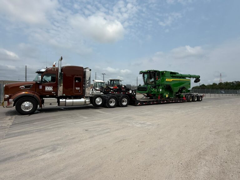 Semi tractor with lowboy trailer parked in yard, carrying green combine harvester and smaller farm equipment.