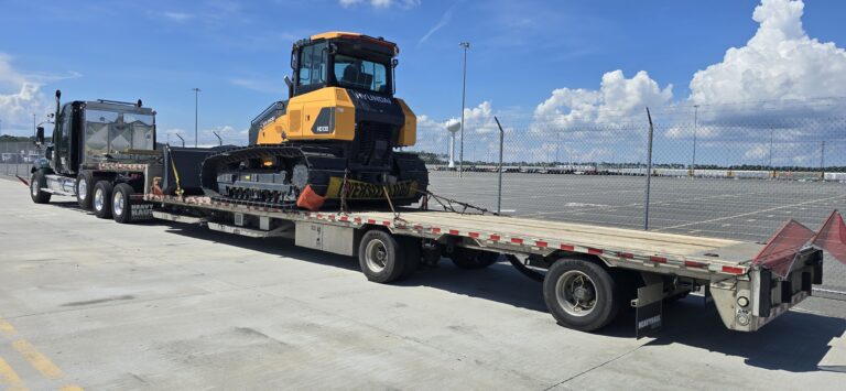 Flatbed trailer parked at a yard with yellow road-construction roller secured on the deck. Support vehicle visible on left.