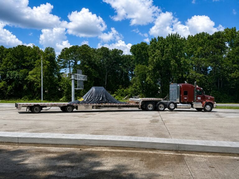 Semi tractor with step deck trailer parked in a paved yard, carrying covered industrial equipment
