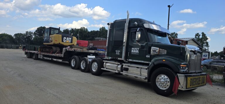 Semi tractor with flatbed trailer carrying a yellow CAT excavator and red shipping container in a yard.