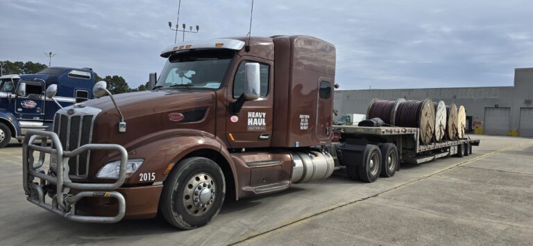 Semi tractor with Heavy Haul Solutions branding parked with loaded step deck trailer carrying cable spools at industrial yard.