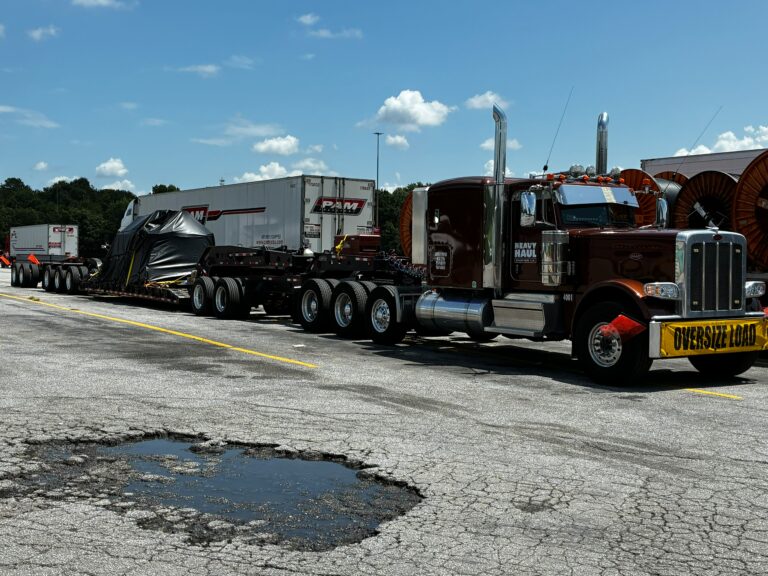 Heavy Haul Solutions semi tractor with lowboy trailer carrying oversized industrial equipment parked in parking lot with oversize load sign visible.