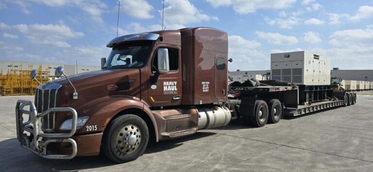 Heavy Haul Solutions semi tractor with RGN trailer parked in yard with industrial equipment and buildings in background.