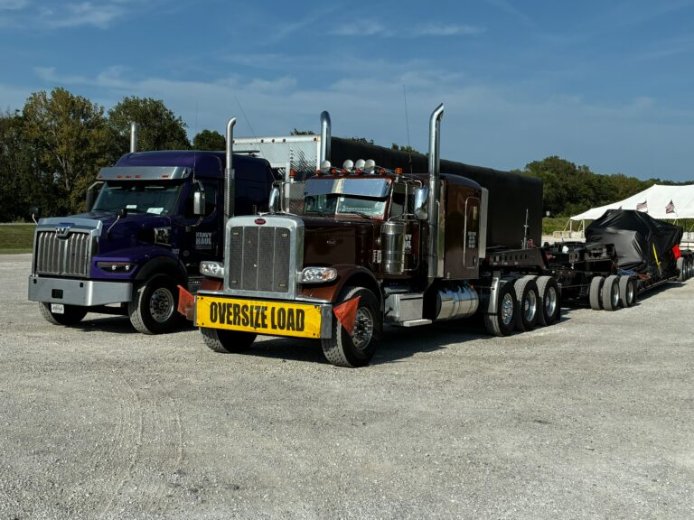 Two heavy-haul tractors parked side-by-side with lowboy trailers carrying oversized industrial equipment, yellow oversize load signs visible.
