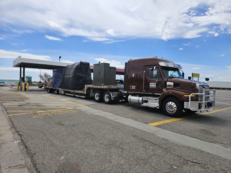 Semi tractor with step deck trailer carrying large industrial equipment parked at terminal with gas station canopy visible in background.