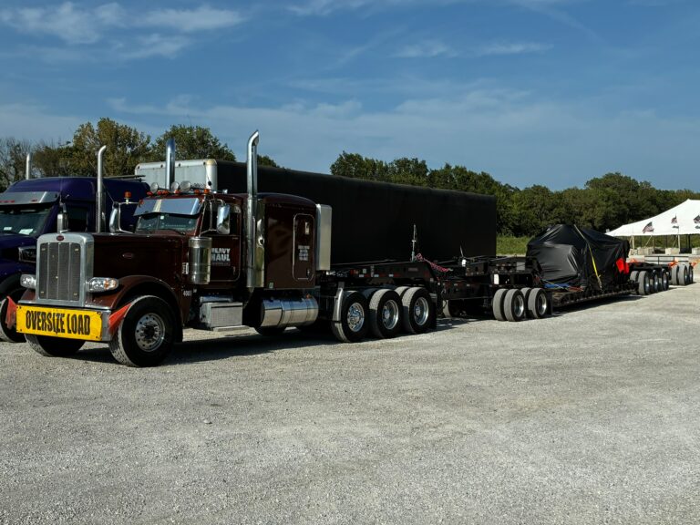 Semi tractor with lowboy trailer carrying oversized black industrial equipment, marked with oversize load sign, parked at facility with tent and trees in background.
