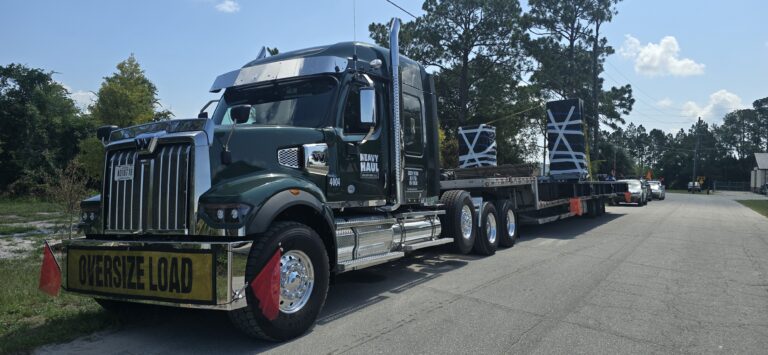Semi truck with flatbed trailer carrying oversized load structures, marked with oversize load signs, parked on residential road.