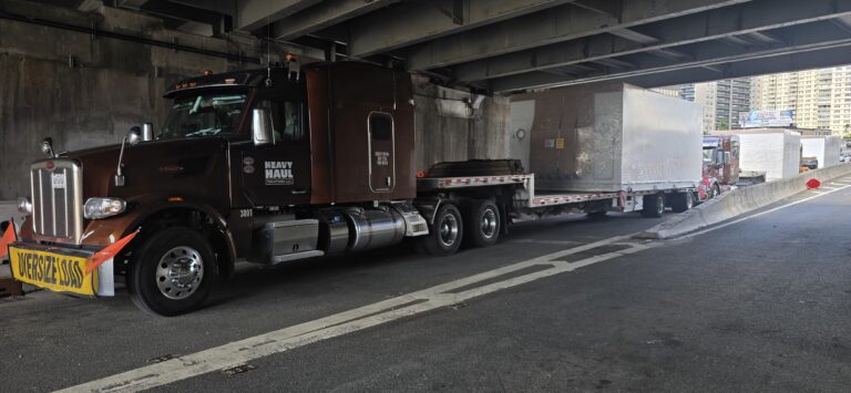 Heavy Haul Solutions tractors with step deck trailer driving under highway overpass in urban area.
