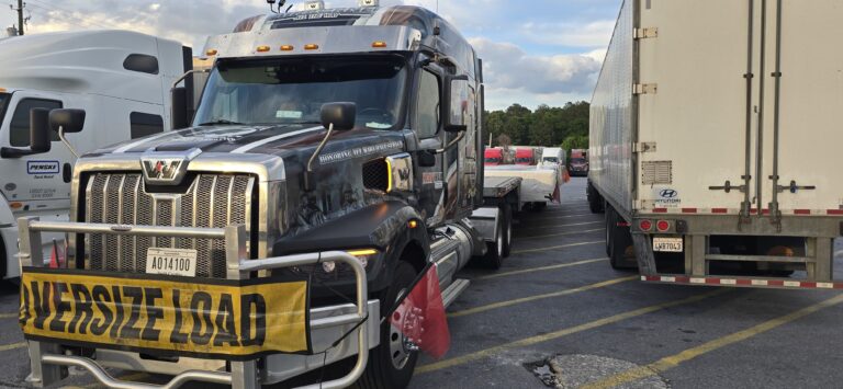 Semi tractor with yellow oversized load banner parked alongside other trucks and trailers at a terminal or staging area.