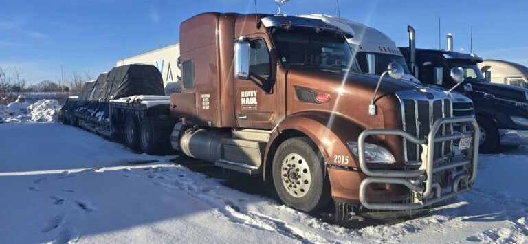 Semi tractor with tarped step deck trailer parked in snowy yard, Heavy Haul Solutions branding visible on cab door.