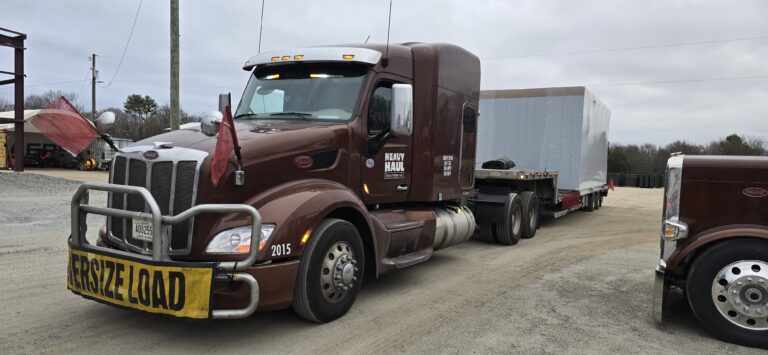 Semi tractor with oversize load banner parked at yard with trailers and equipment visible in background