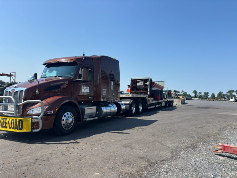 Semi tractor with step deck trailer parked in yard, with oversize load secured on deck and other equipment nearby.