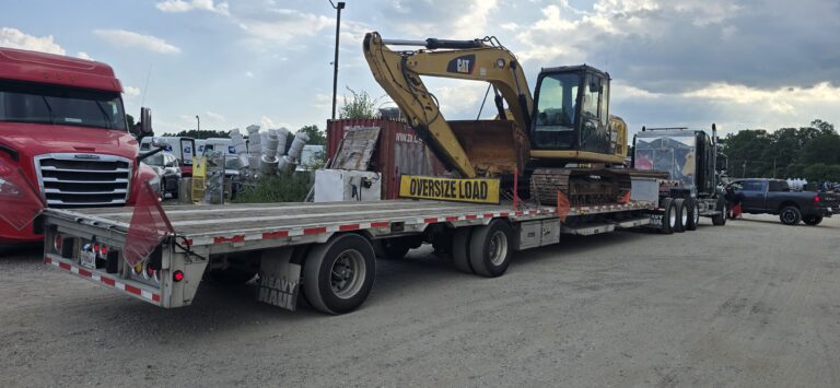 Flatbed trailer carrying yellow CAT excavator with oversize load banner, parked at yard with other trucks and equipment.