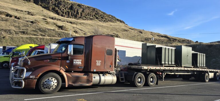 Heavy Haul Solutions semi tractor with flatbed trailer carrying large industrial transformers parked at roadside with hillside in background