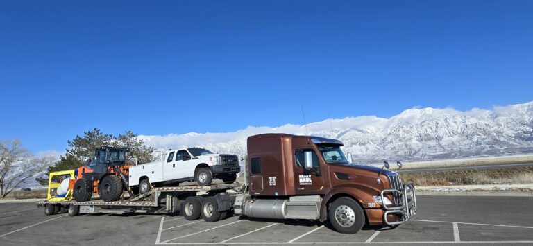 Semi tractor with brown cab towing step deck trailer carrying wheel loader and white pickup truck in parking lot with snowy mountains in background