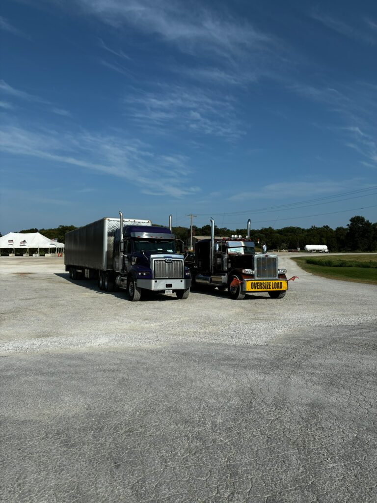Two semi trucks parked on a lot, one with an oversized load banner, under blue sky. And the other hauling a Conestoga trailer.