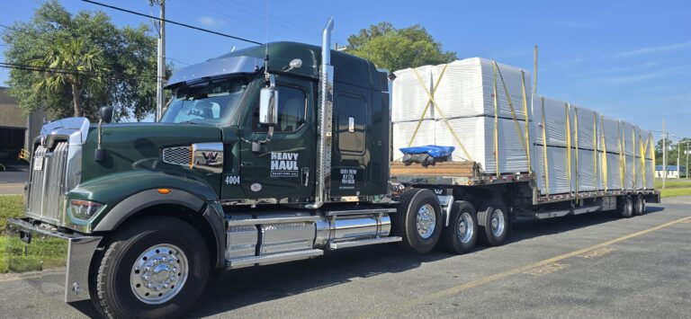 Semi tractor with step deck trailer carrying wrapped cargo parked on roadside with trees and utility poles in background.