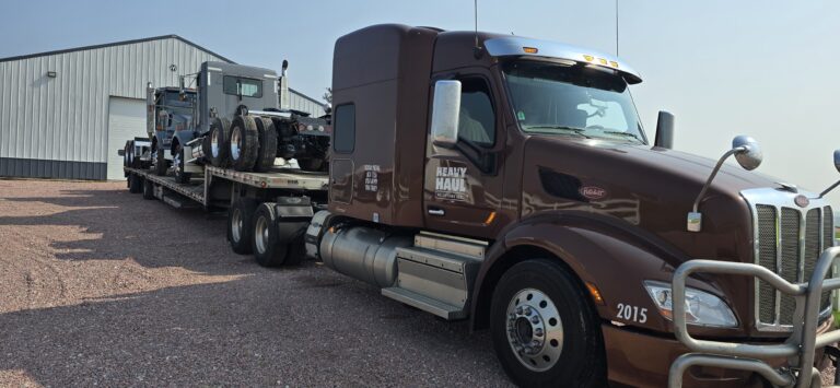 Semi tractor with step deck trailer loaded with grey tractors parked at a yard next to a shop.