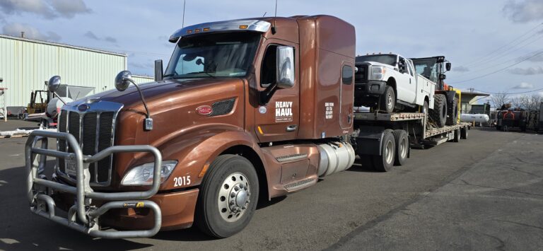 Heavy Haul Solutions semi tractor with brown cab parked at yard with white service truck and machinery on step deck trailer.
