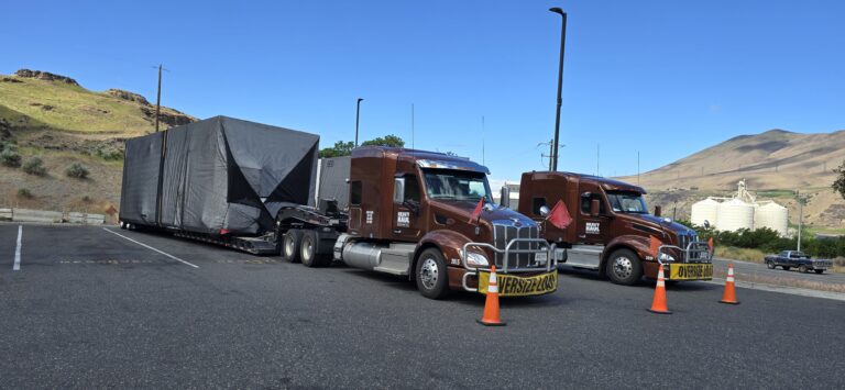 Two Heavy Haul Solutions semi tractors parked and hauling oversized modular buildings, hills in background.