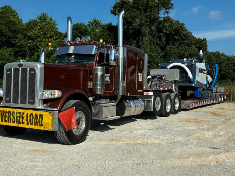Semi tractor with oversized load sign parked on lot with lowboy trailer carrying industrial equipment and machinery.