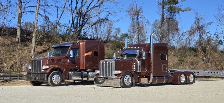 Two brown semi tractors parked side by side on a gravel lot with bare trees in the background. Rust-bucket truck