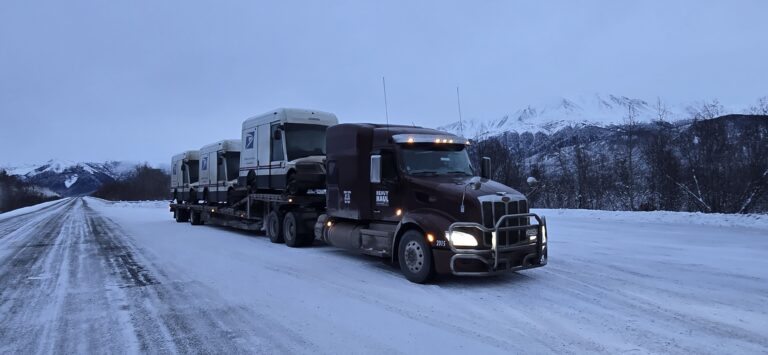 Heavy Haul Solutions tractor hauling Oshkosh NGDVs mail tri snowy road with snow-covered mountains in background