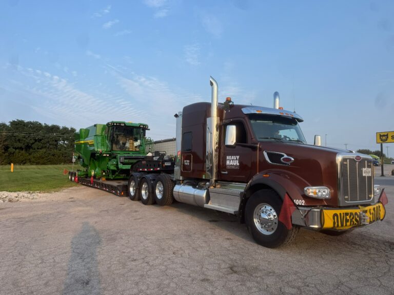 Semi tractor with maroon cab and chrome stacks hauling RGN trailer carrying green combine harvester.