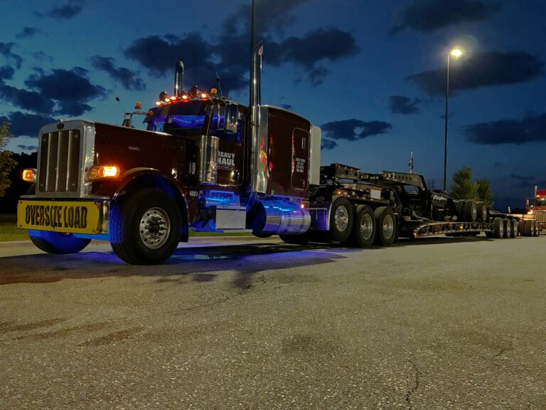 Semi tractor with blue accent lighting and heavy-haul trailer parked at night, yellow 'Oversize Load' sign visible on front fender.