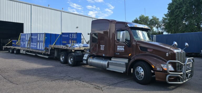 Semi tractor with step deck trailer carrying blue industrial units and pallets at a yard with industrial buildings and trees in background.