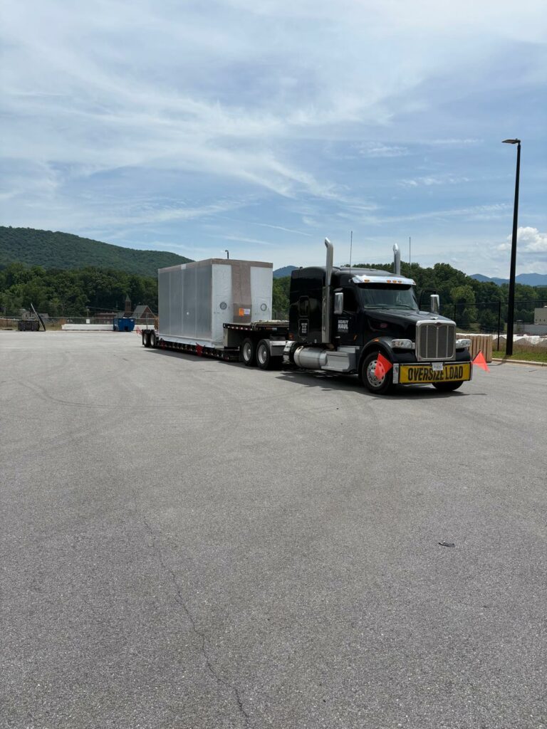 Semi tractor with oversize load banner parked in yard hauling RGN trailer, mountains and trees in background.